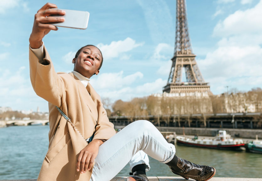 junge Frau sitzt auf Brücke über der Seine und macht ein Selfie mit Eiffelturm im Hintergrund