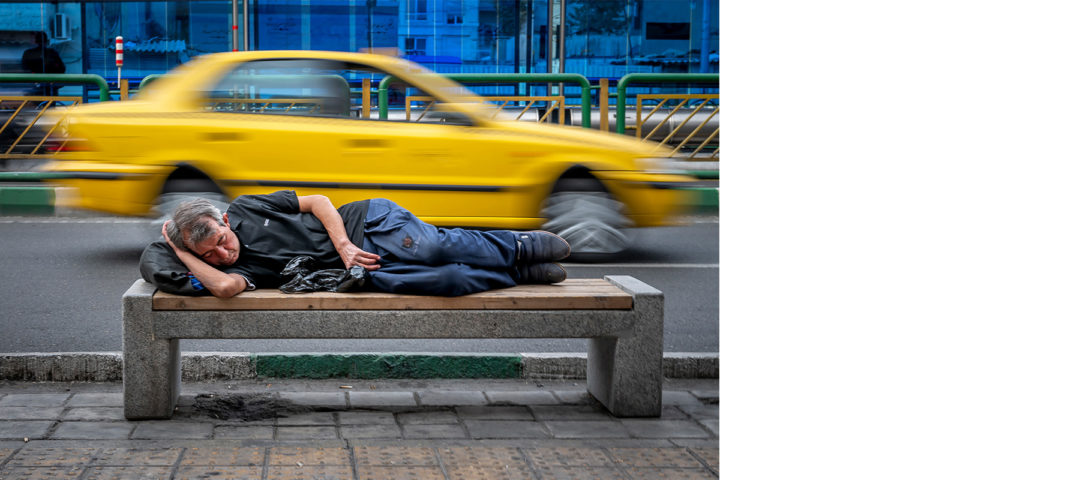 Mann schläft auf einer Parkbank. In Hintergrund fährt ein Taxi dabei.