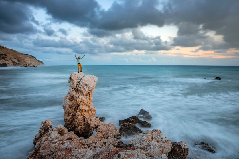 Ein Mann steht auf einem Felsen an der Küste. Die Wellen des Meeres sind aufgrund der langen Belichtungszeit verwischt.