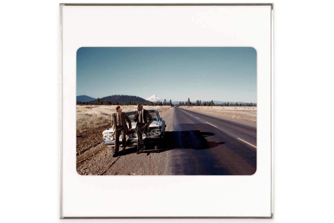 Zwei Männer stehen an einen silbernen Pontiac gelehnt am Rand einer Straße, die im Hintergrund in weite Ferne führt.